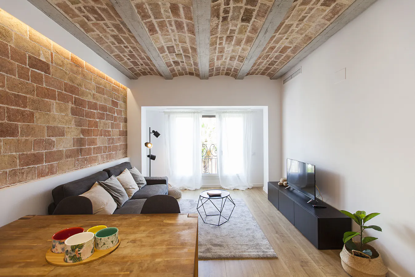 Living room with brick wall, exposed brick ceiling, gray sofa, TV, and wooden table with colorful bowls. Balcony with white curtains.