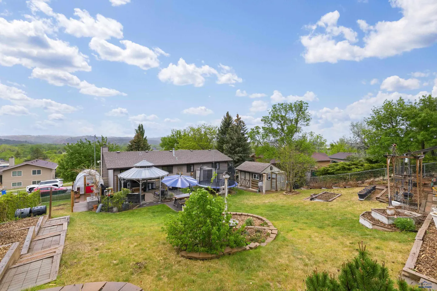 Backyard of a suburban home with a gazebo, trampoline, small shed, garden beds, and a grassy lawn under a blue sky.