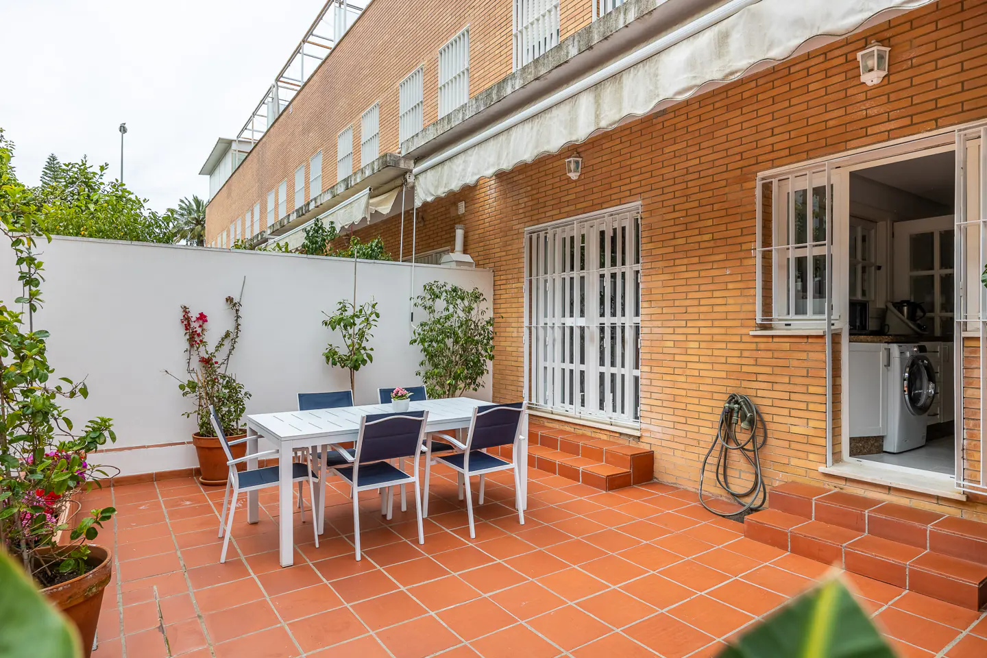 Outdoor patio with terracotta tiles, a white table with six blue chairs, and a brick building in the background. Plants in pots add greenery.