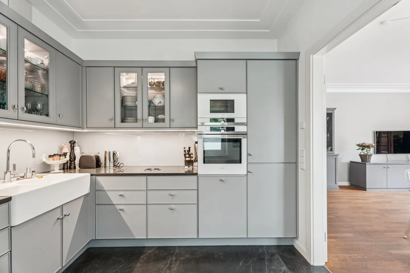 Modern kitchen with gray cabinets, black countertops, and a white farmhouse sink. Glass-front cabinets display dishware. Oven and microwave are built-in.