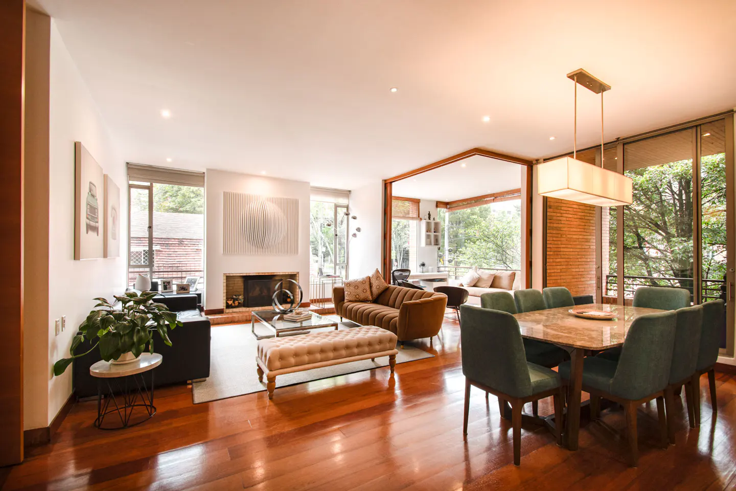 Open-concept living and dining room with hardwood floors, brown sofa, fireplace, and green dining chairs. Large windows with trees outside.