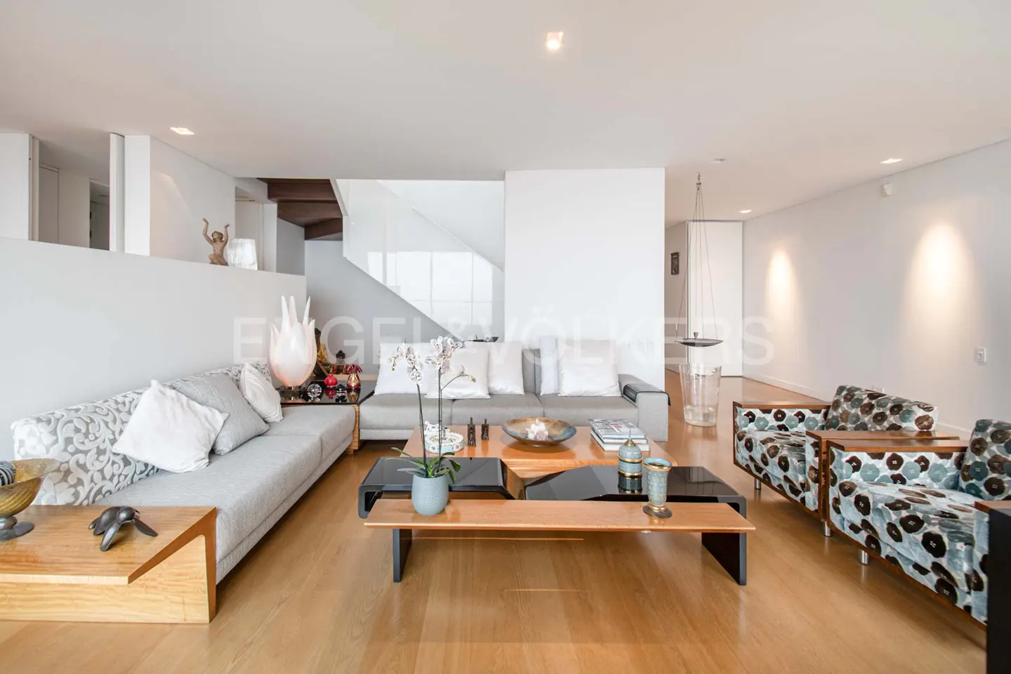 Bright, modern living room with wood floors, gray sofas, patterned armchairs, and a unique coffee table. White walls and staircase in the background.