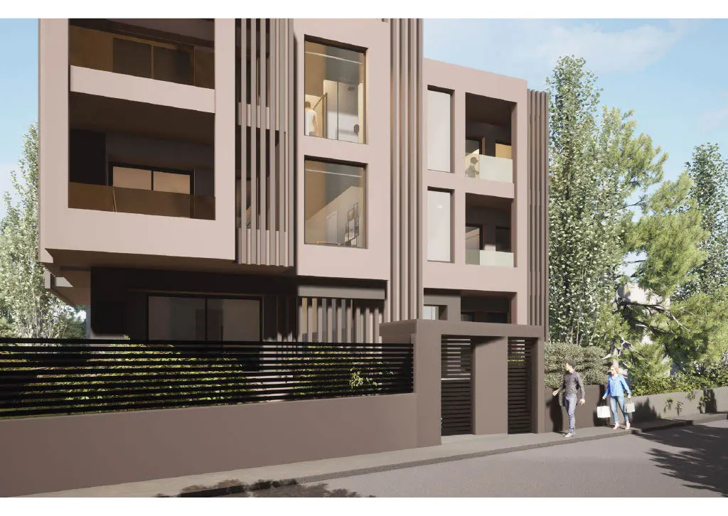 Modern three-story apartment building with balconies, a dark fence, and two people walking on the sidewalk.