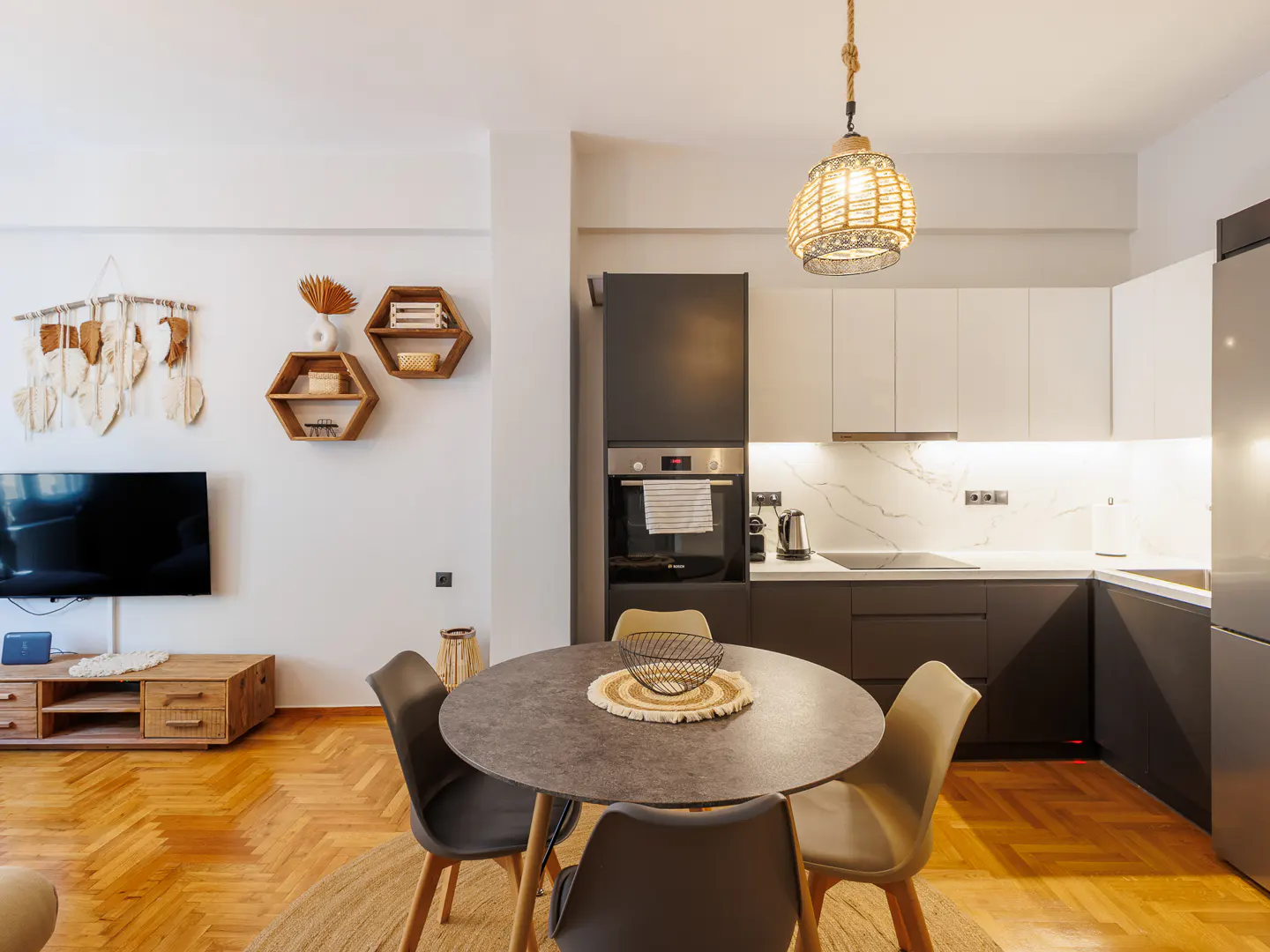 A modern apartment interior with a round dining table, chairs, and a kitchen with white and gray cabinets. A TV and decorative shelves are on the wall.