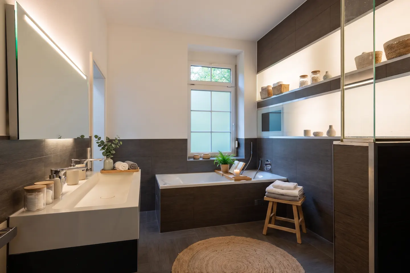 Bright bathroom with a white sink, a tub with a wooden tray, and a round rug. Shelves with decorative items are on the right.