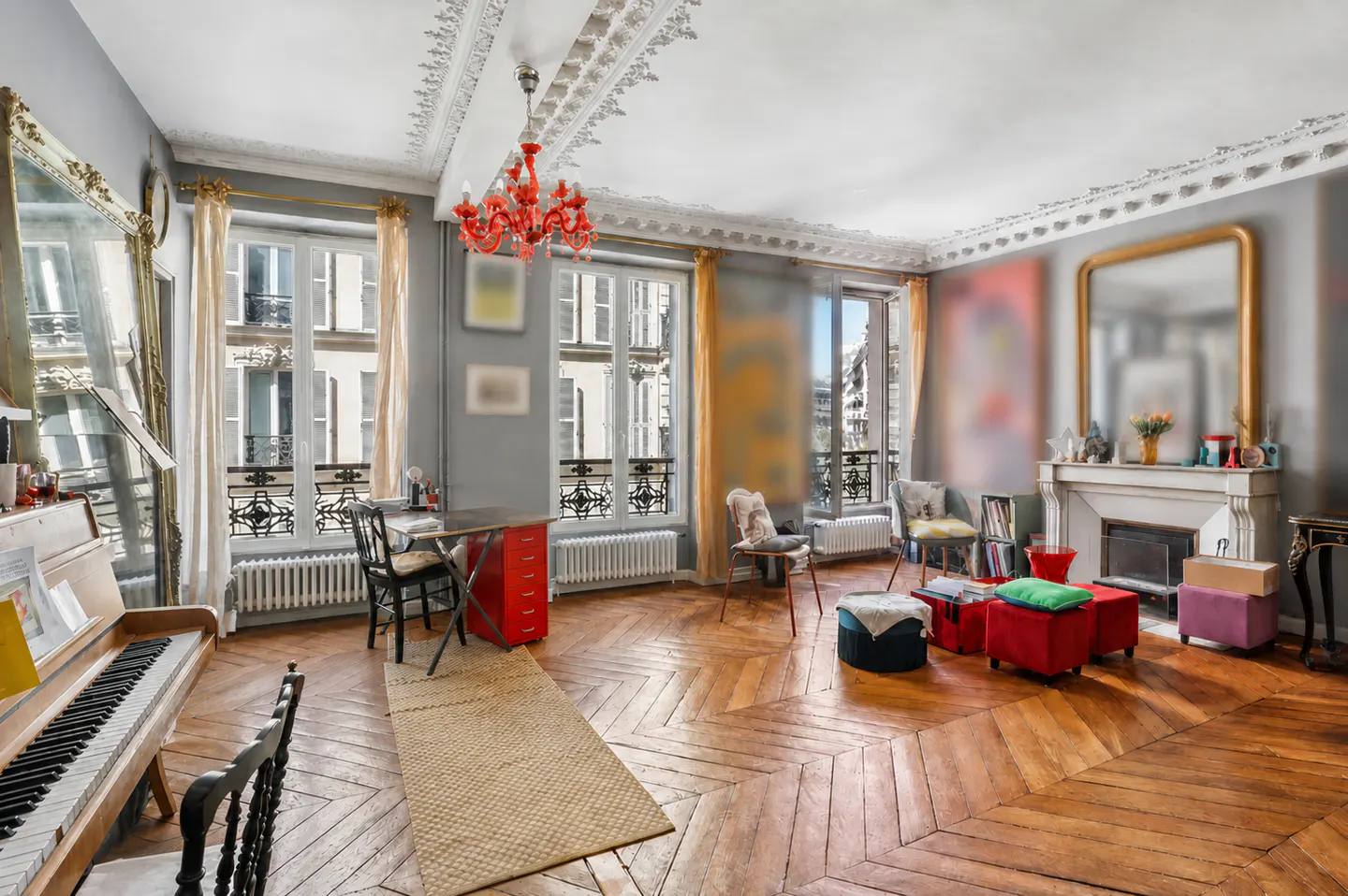 Bright living room with herringbone wood floors, grey walls, and large windows. A red chandelier hangs from the ornate ceiling. Piano and fireplace are visible.