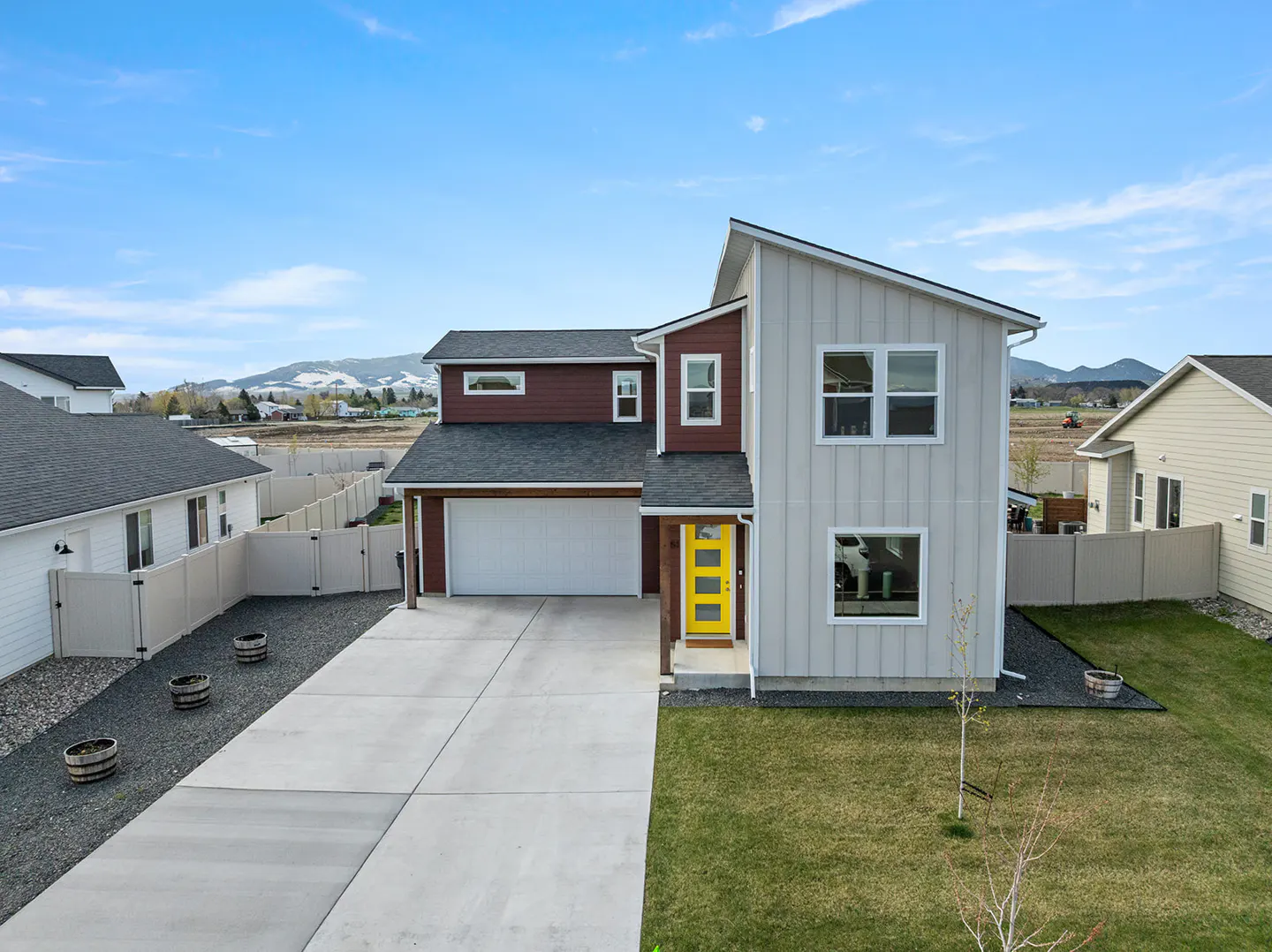 Two-story house with gray and maroon siding, a white garage door, and a bright yellow front door. Green lawn and blue sky.