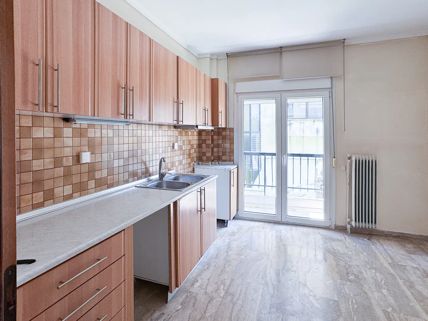 Bright kitchen with wood cabinets, marble floors, and a double sink. A window overlooks a balcony.