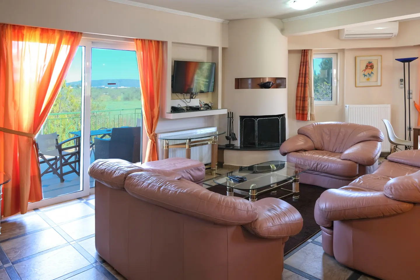 Living room with pink leather sofas, fireplace, TV, and balcony with orange curtains overlooking a green landscape.
