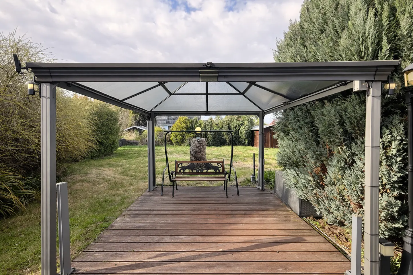 A backyard gazebo with a swing bench on a wooden deck, surrounded by green grass and trees.