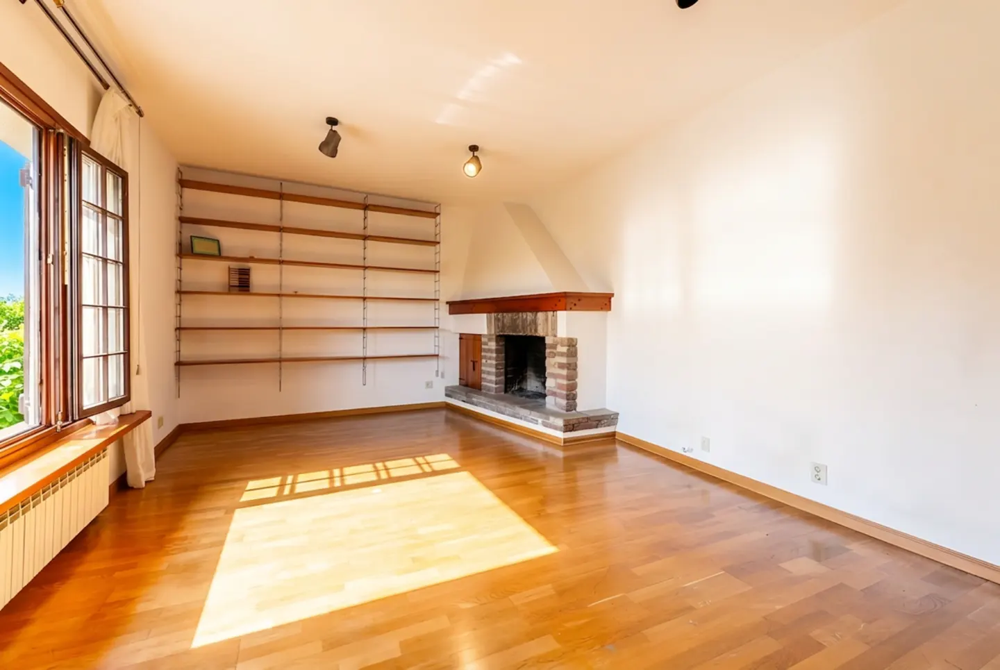 Bright, empty living room with hardwood floors, white walls, and a brick fireplace. A large window and built-in shelves add character.