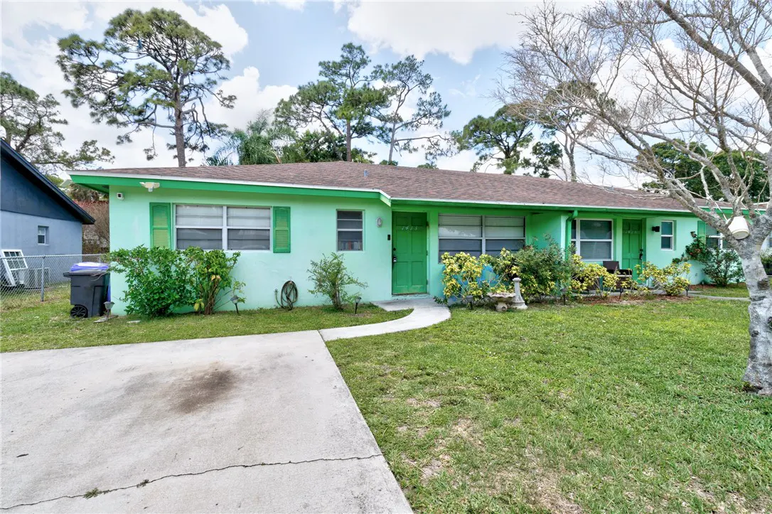 Exterior view of a light green, single-story duplex with green shutters and doors, a concrete driveway, and a grassy lawn.