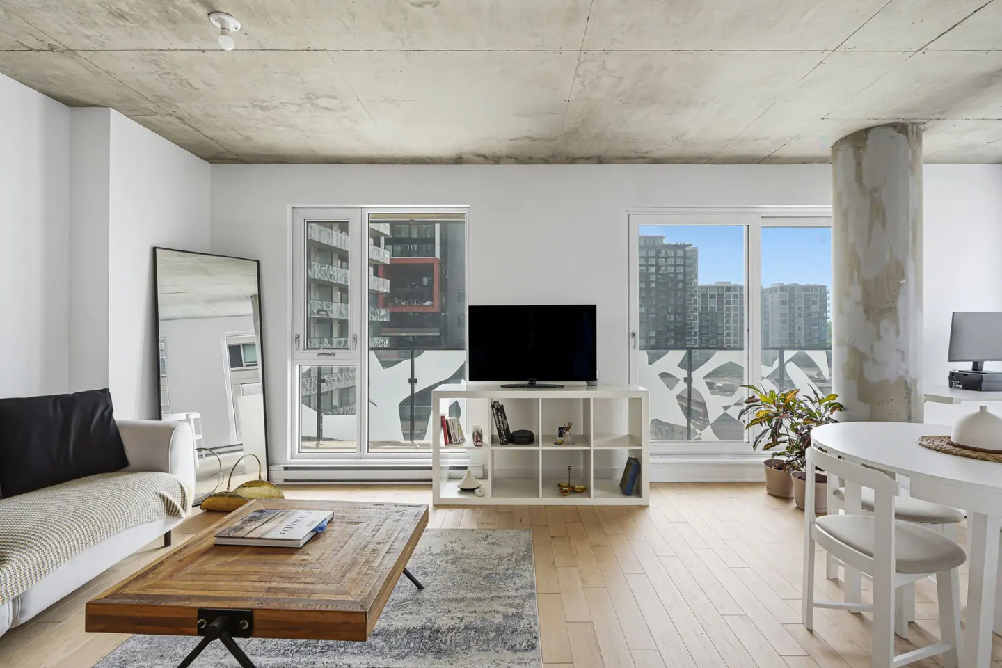 Modern living room with concrete ceiling, white walls, wood floors, TV on a white shelf, and large windows with city views.