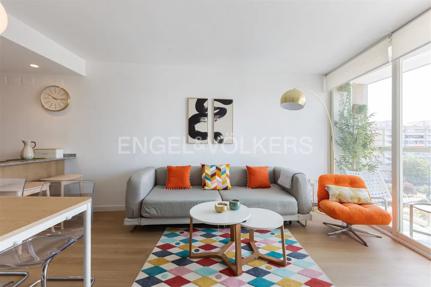 Bright living room with gray sofa, orange chair, and colorful rug. Large windows offer city views. Modern decor with art and gold lamp.
