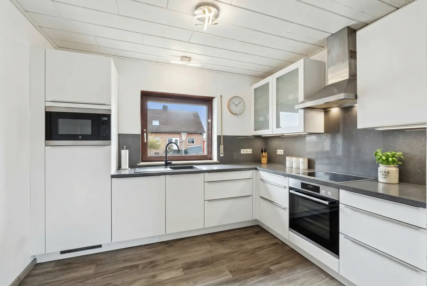 A modern kitchen with white cabinets, gray countertops, and wood-look flooring. A window overlooks a house.