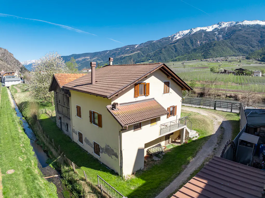 Exterior view of a two-story house with a brown roof, beige walls, and brown shutters, set against a backdrop of mountains and a blue sky.