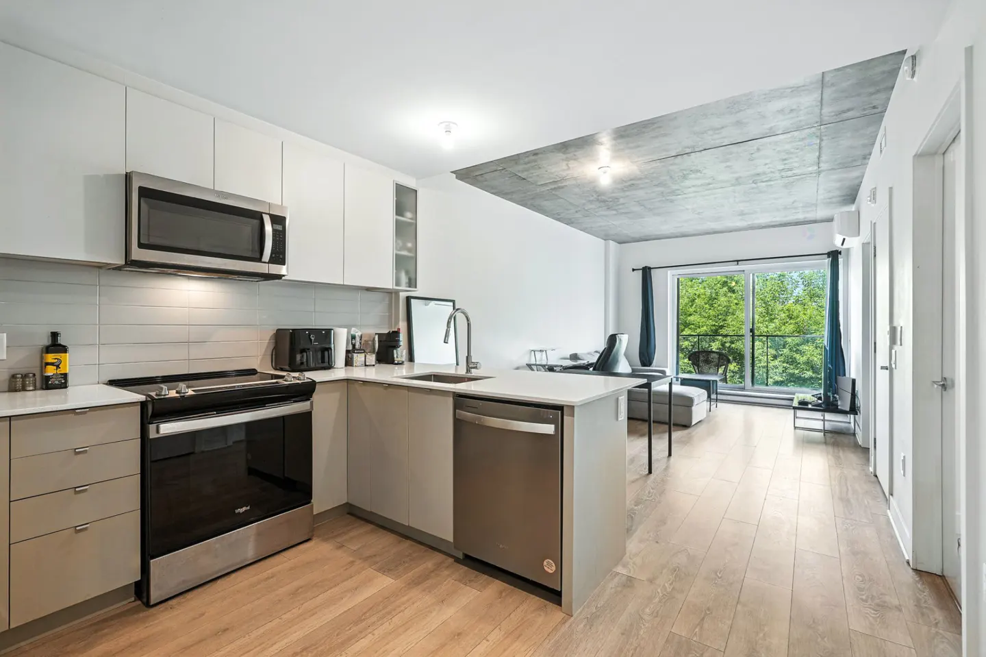 A modern apartment interior with white walls, light wood floors, and a kitchen with stainless steel appliances. A balcony with green trees is visible.