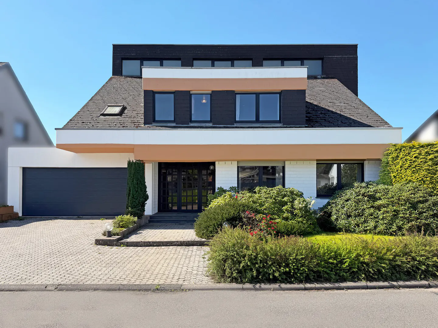 Modern two-story house with a black garage door, dark roof, and well-maintained landscaping. The house has a white and tan facade.