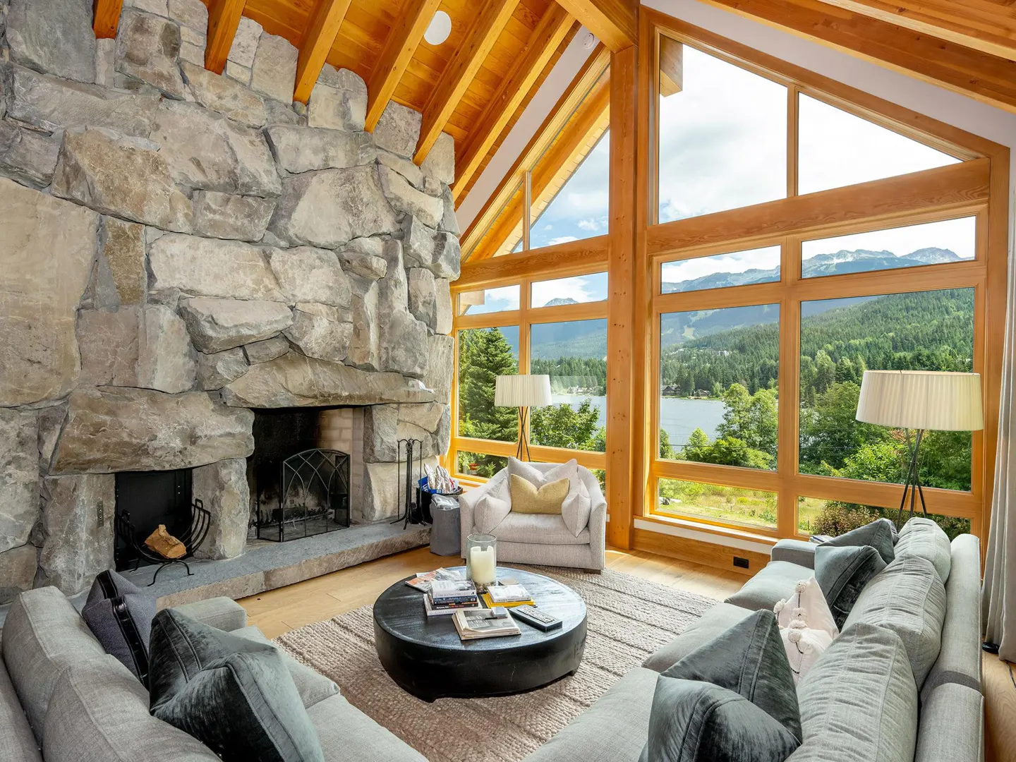 Living room with stone fireplace, wood ceiling, and large windows with lake and mountain views. Gray sofa and round coffee table.