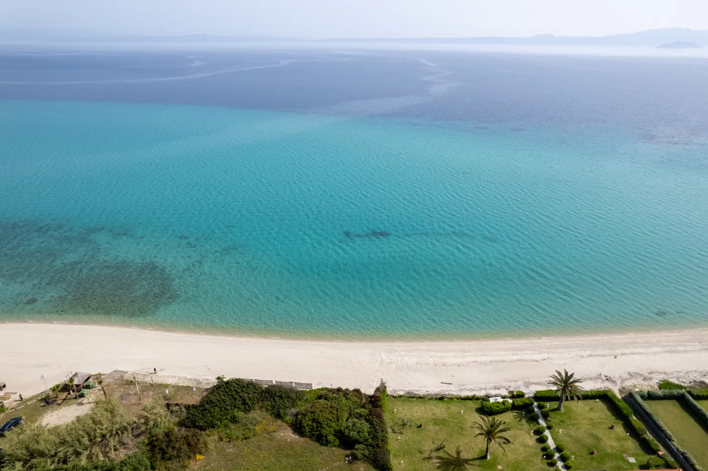Aerial view of a sandy beach meeting turquoise water, with green lawns and palm trees in the foreground.
