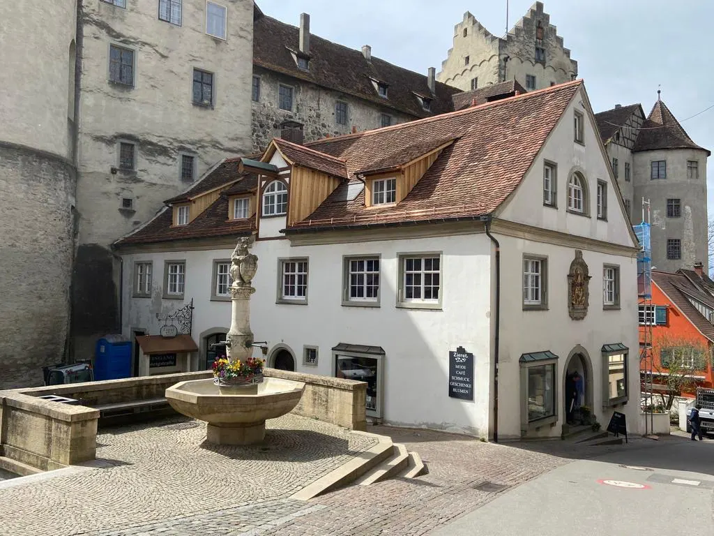 Exterior view of a white building with a red tile roof, a stone fountain, and a castle in the background.