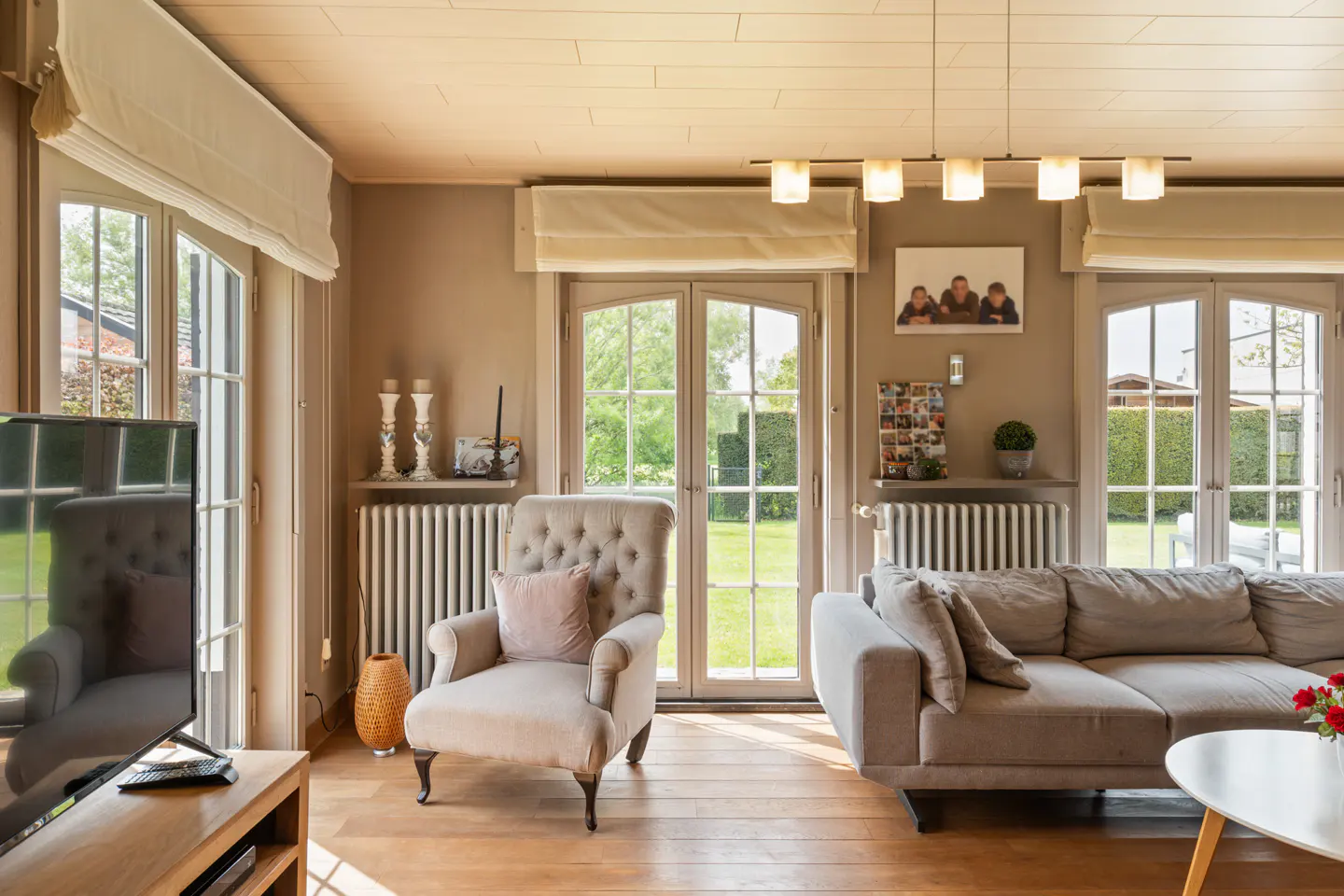 Living room with hardwood floors, beige walls, and natural light. A tufted armchair and gray sofa face French doors to a green yard.
