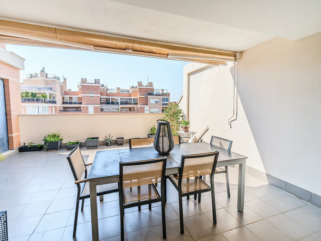 Spacious apartment terrace with a dining table, chairs, potted plants, and a view of neighboring brick buildings.