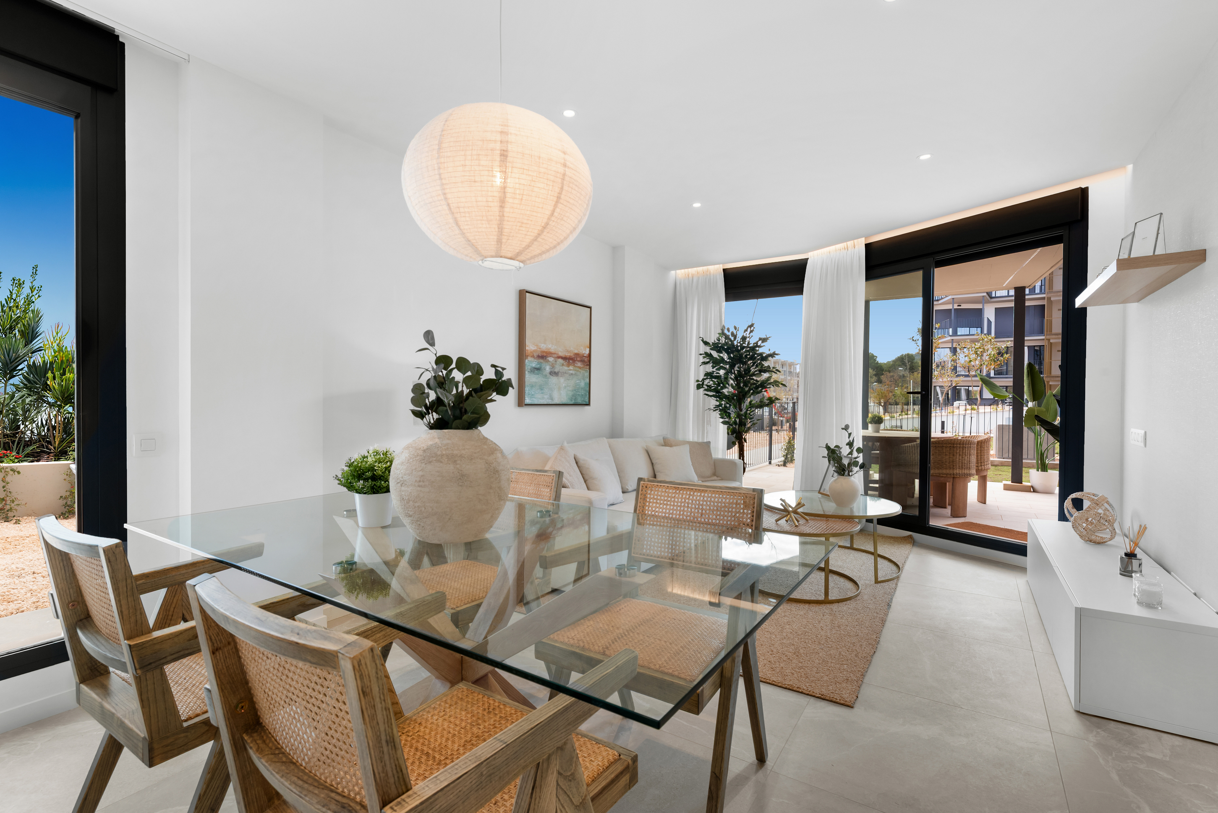 Bright, modern living space with glass table, woven chairs, and white walls. Large windows offer outdoor views. A white sofa and decor add to the airy feel.