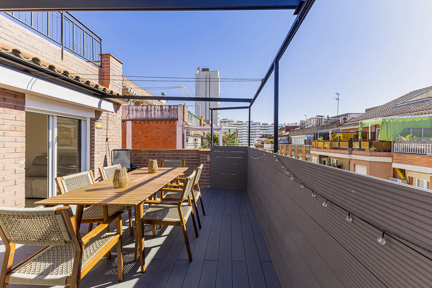Outdoor patio with a wooden table, chairs, and string lights. Brick buildings and a blue sky are in the background.