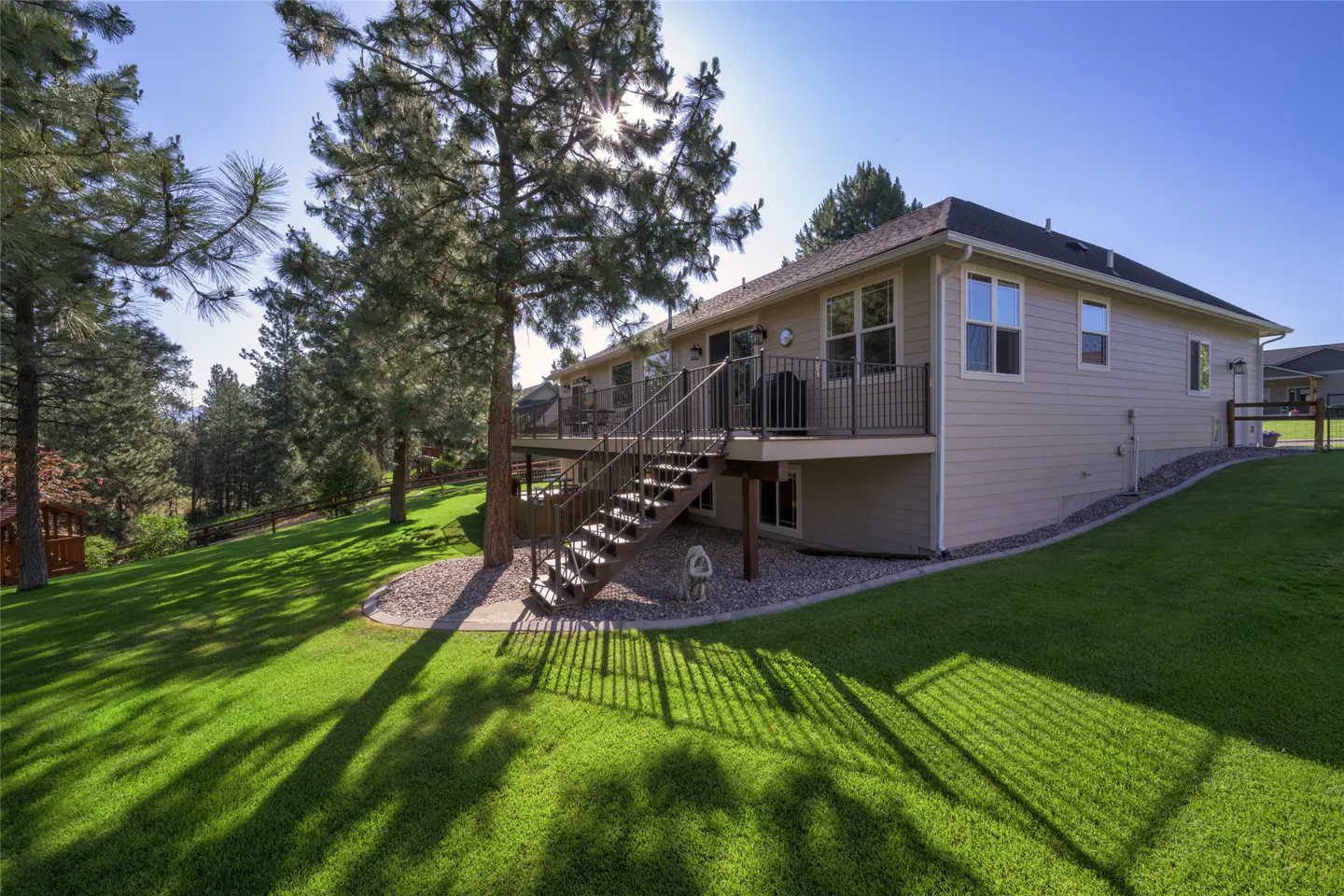 Tan house with a deck and stairs leading to a green lawn. Trees provide shade, and the sky is blue.