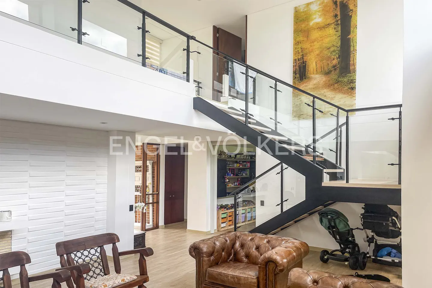 Bright, modern living room with brown leather chairs, wooden chairs, and a black staircase with glass railings. A large autumn forest print hangs on the wall.