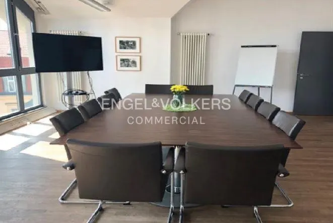 A modern conference room with a large wooden table, black chairs, a wall-mounted TV, a whiteboard, and yellow flowers.