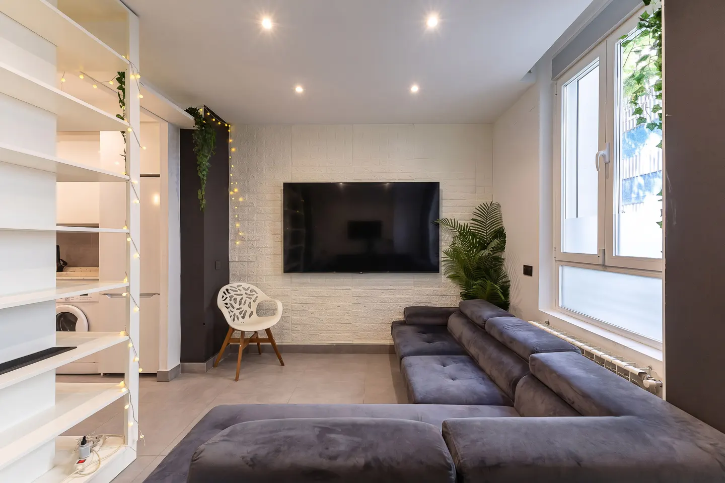 Living room with gray sectional sofa, TV on white brick wall, white chair, and window with plants.
