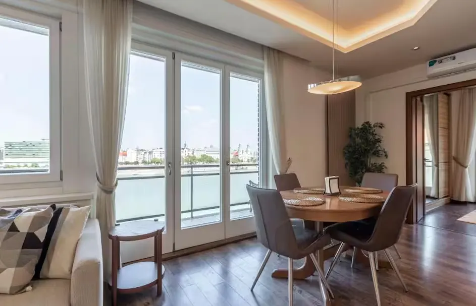 Modern dining area with a round wooden table, four grey chairs, and large glass doors overlooking a river and city skyline.
