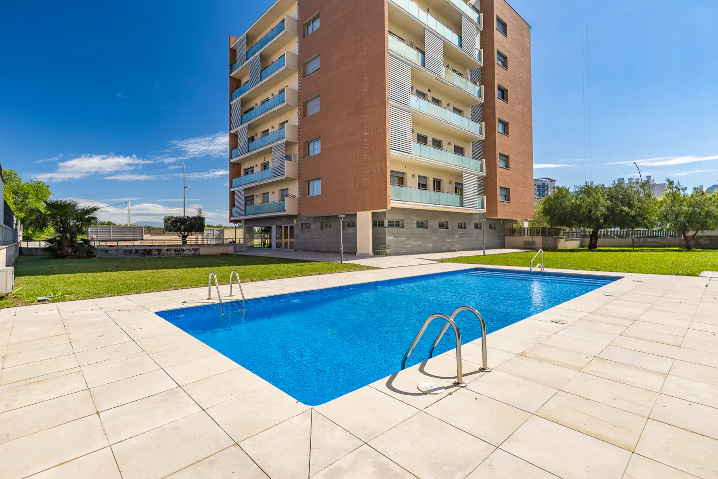 Outdoor pool with metal ladders, surrounded by a tiled patio and green lawn, in front of a multi-story brick apartment building under a blue sky.