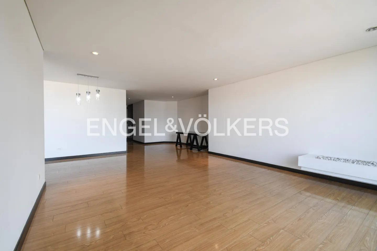 Bright, empty room with light wood floors and white walls. A black table sits in the background. Engel & Völkers logo is superimposed.
