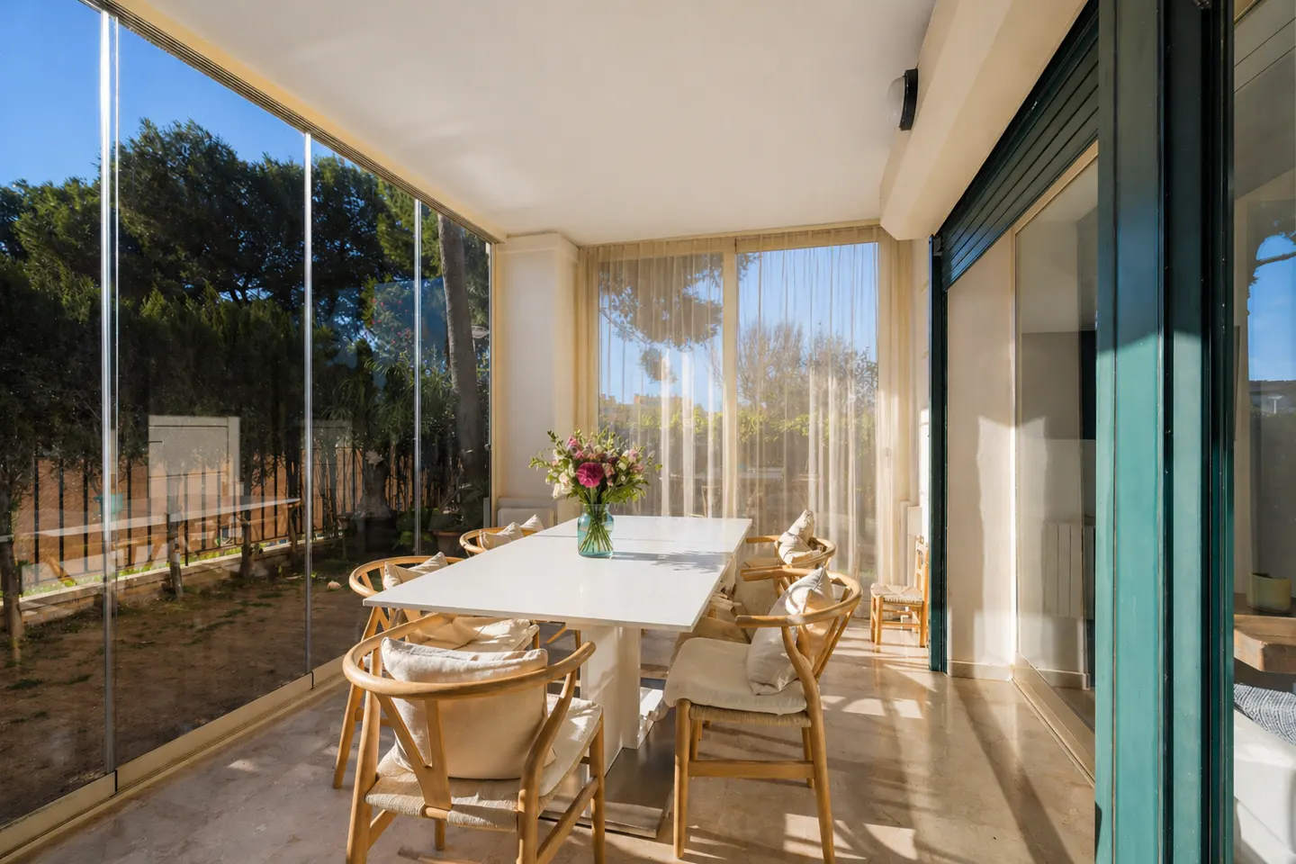 Bright sunroom with a white table, wooden chairs with cushions, and a vase of flowers. Glass walls offer a view of trees and sky.