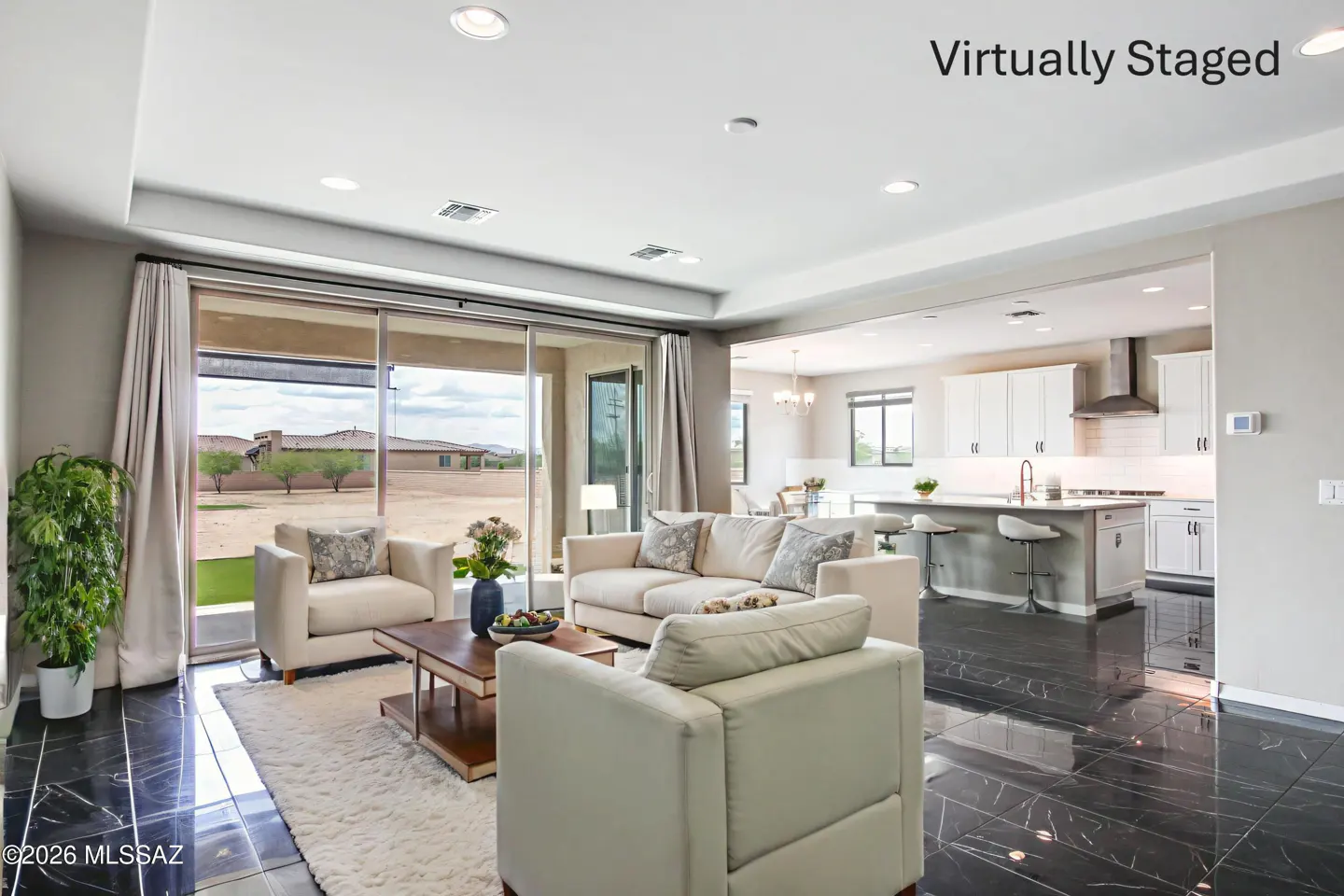 Bright, virtually staged living room with white furniture, black marble floors, and an open floor plan leading to a white kitchen. Large sliding glass doors offer a view of the desert landscape.