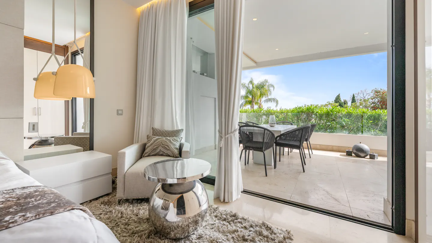 Bedroom view to patio with table and chairs through sliding glass doors with white curtains. Armchair and silver table in foreground.