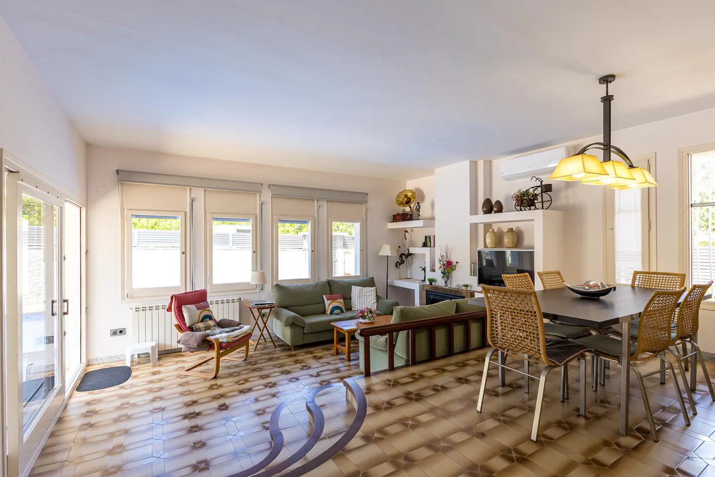 Bright living and dining area with green sofas, a dark wood table, and patterned tile flooring. Natural light streams through multiple windows.