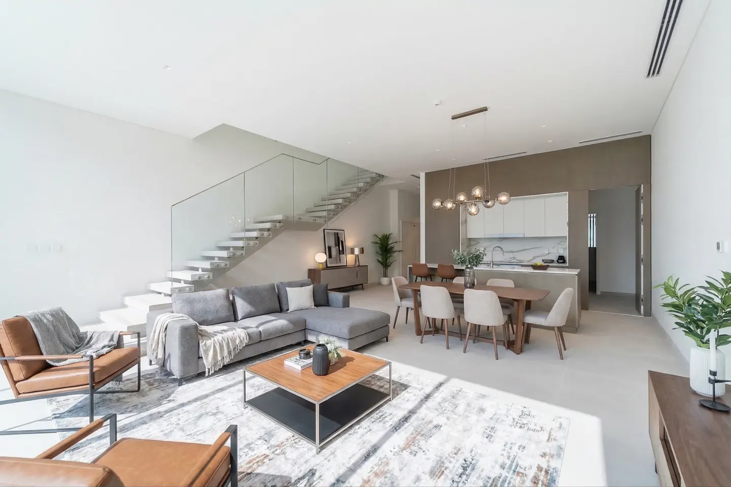 Bright, modern living space with gray sectional, wood table, and glass-railed stairs. Kitchen and dining area visible.