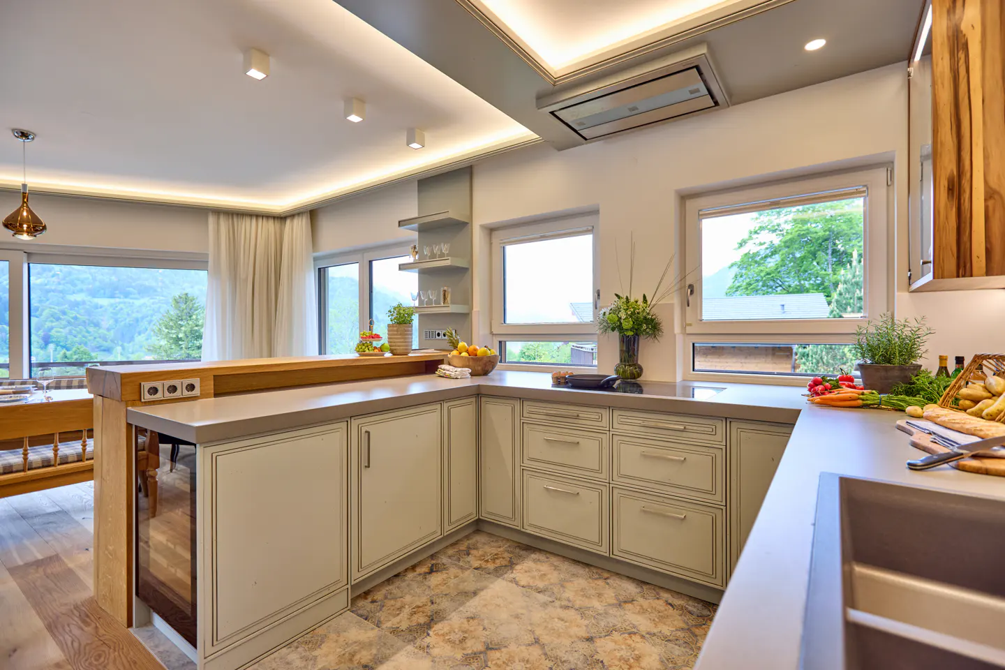 Bright kitchen with gray cabinets, stone floors, and a wood breakfast bar. Windows overlook a green, mountainous landscape.