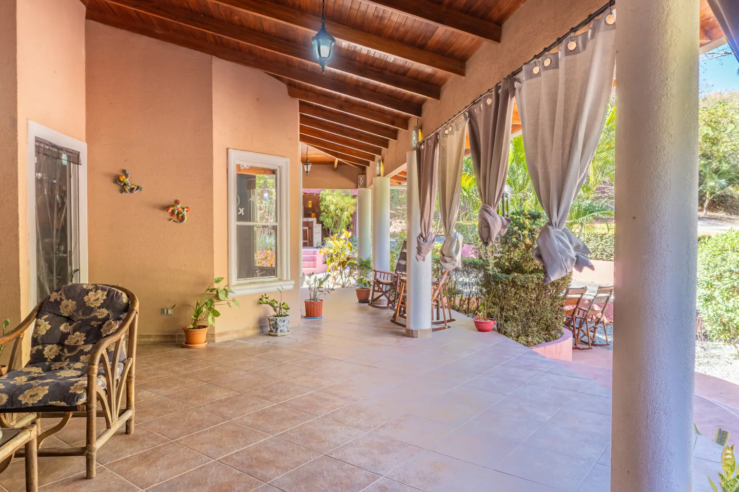 Covered patio with tile floor, peach walls, and wood ceiling. White columns support the roof, and gray curtains hang between them. Rattan furniture and potted plants decorate the space.