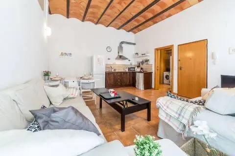 Bright apartment interior with white sofas, a black coffee table, and a kitchen area with brown cabinets. The ceiling has exposed terracotta beams.
