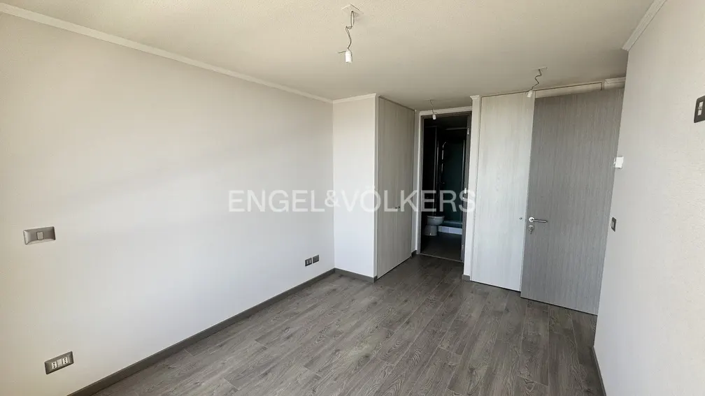 An empty bedroom with gray wood floors, white walls, and a bathroom visible through an open door.