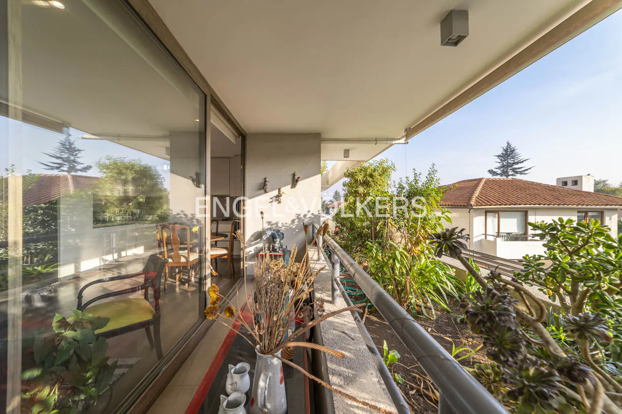 Balcony view with chairs, plants, and a table with vases. Outside, there's a house with a red tile roof and greenery.