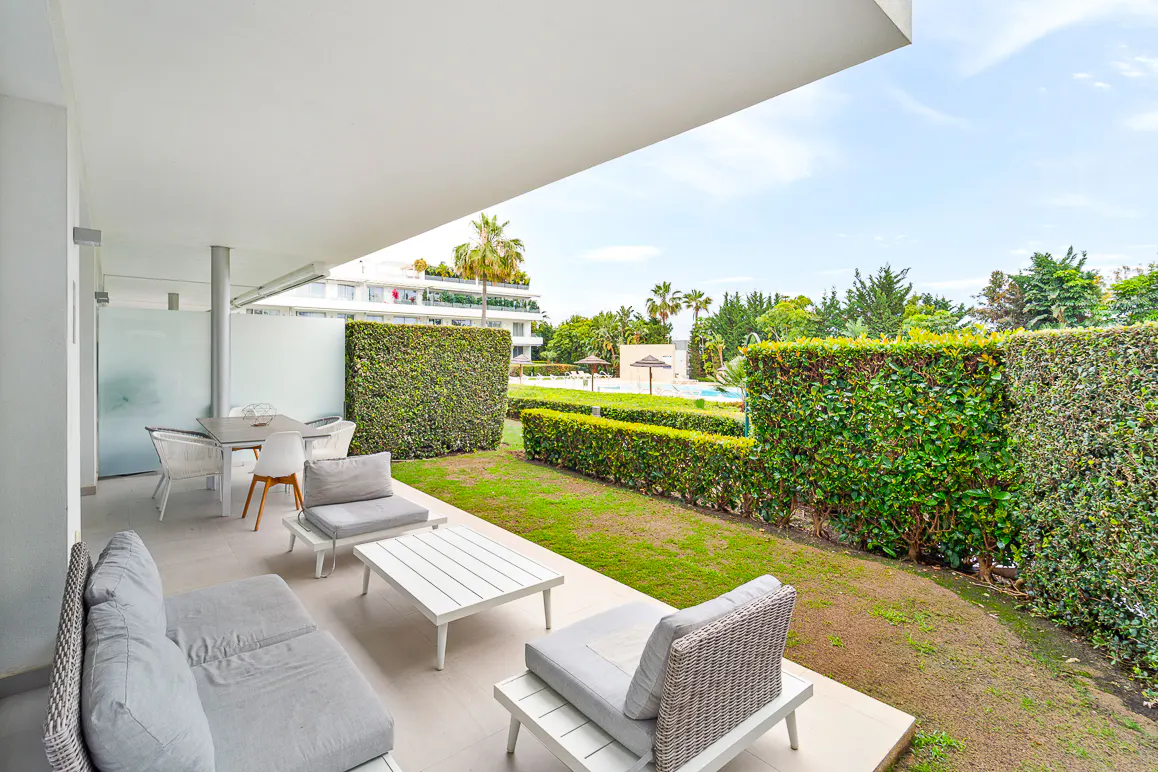 A covered patio with white furniture overlooks a green lawn and trimmed hedges. A pool and building are visible in the background.