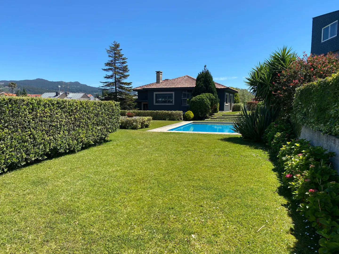 A house with a red tile roof and a blue pool is surrounded by green grass and hedges on a sunny day.