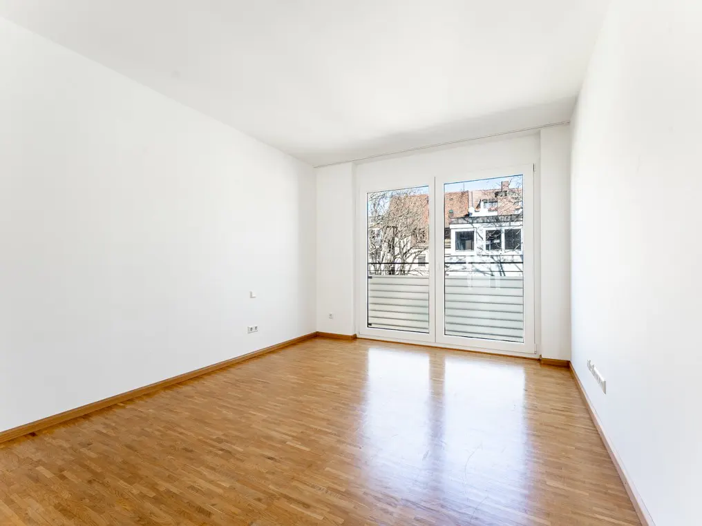 Empty room with white walls, wood floor, and a large window showing a tree and buildings.