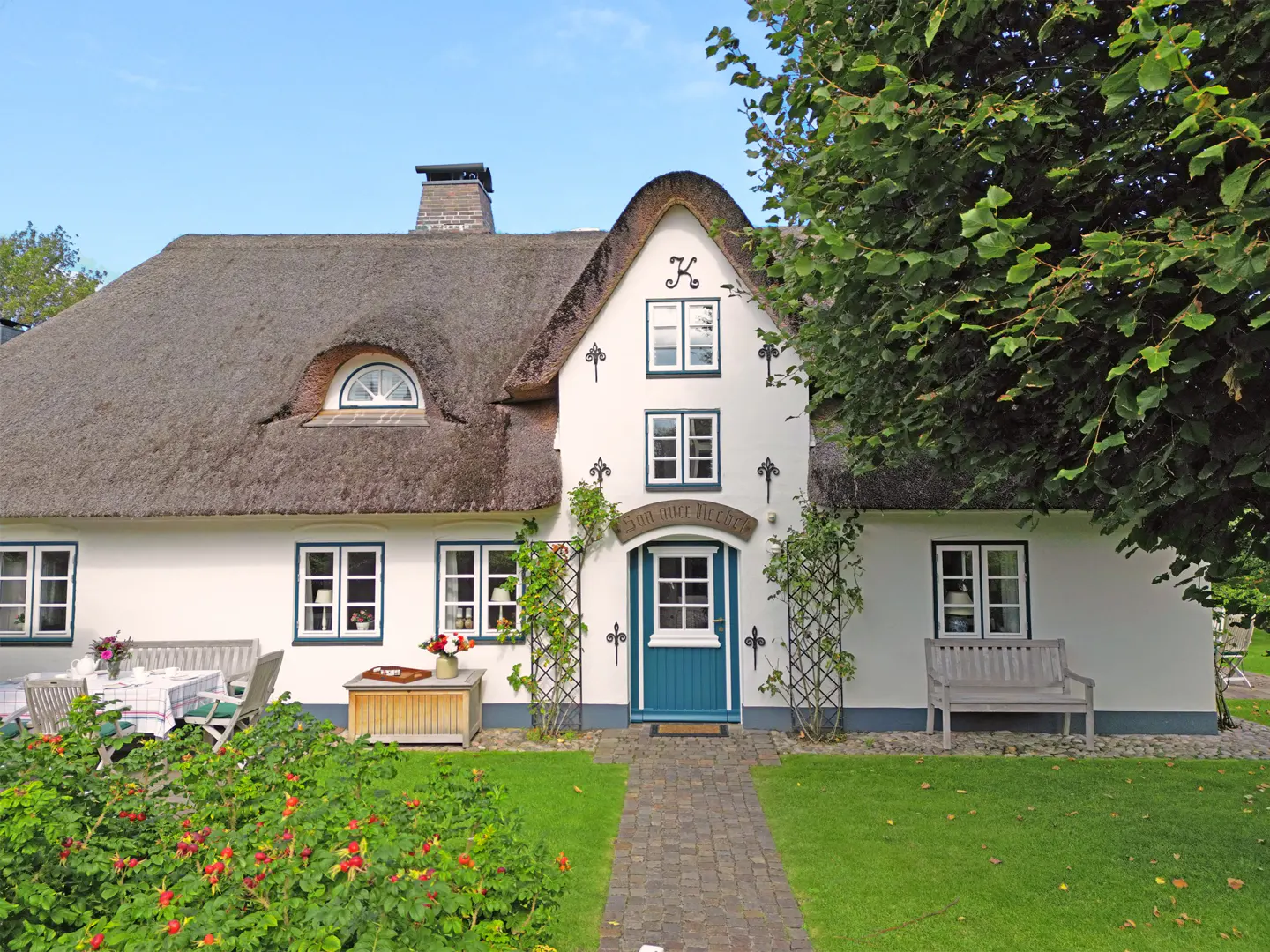 Charming white house with a thatched roof, blue door, and lush green lawn and garden. Outdoor seating visible.